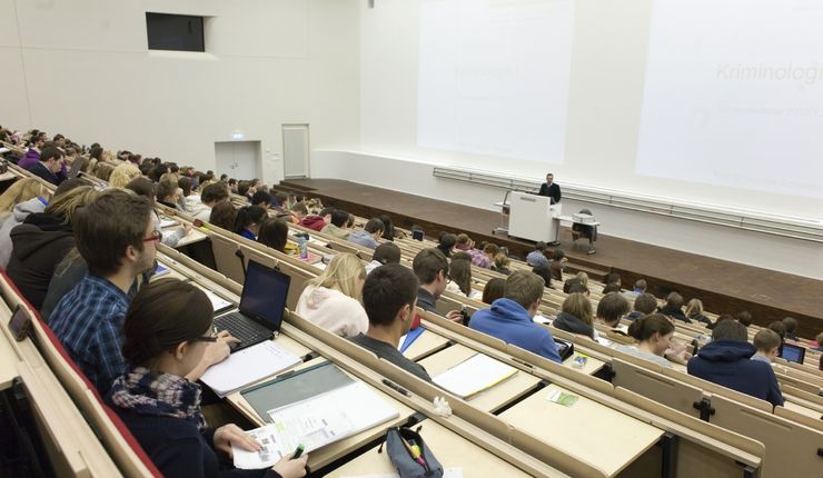 Foto: Vorlesung im vollbesetzten Audimax, am Rednerpult steht ein Professor