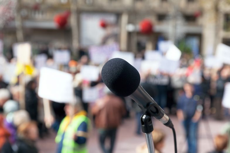 Symbolbild, Mikrofon vor einer unscharfen Menschenansammlung mit Schildern und Transparenten