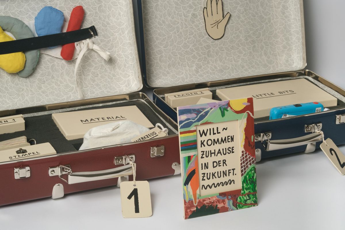 enlarge the image: You can see two open suitcases with numbers on their holders. Inside the suitcases are various envelopes labelled ‘Material’, ‘Little Boxes’ and ‘Stamp’. In front of the suitcases is a colourfully painted picture frame. Inside is the text ‘Welcome home to the future.’