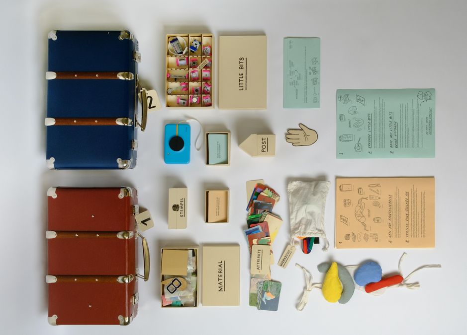 enlarge the image: You can see two open suitcases with numbers on their holders. Inside the suitcases are various envelopes labelled ‘Material’, ‘Little Boxes’ and ‘Stamp’. In front of the suitcases is a colourfully painted picture frame. Inside is the text ‘Welcome home to the future.’