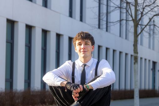 Noah Dejanovic sitzt im Außenbereich des Campus Jahnallee auf einem Stein und schaut lächelnd in die Kamera.