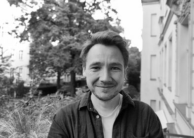 Schwarz-Weiß-Foto von Christian Rau, im Hintergrund ein Teil eines Wohnhauses und ein Hinterhof