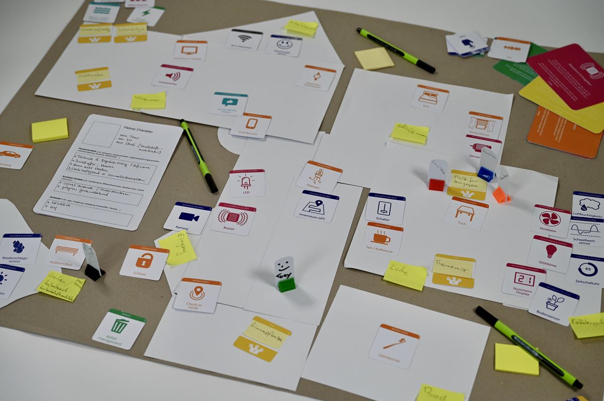 enlarge the image: You can see a brown cardboard box. On it are white sheets of paper in various formats. There are game figures on them. Colourful cards are scattered around the edge of the box. There are also pens and post-its on the box.