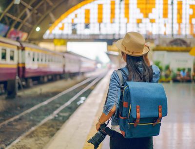 Aufnahme einer Bahnhofshalle. Links sind Gleise zu sehen sowie ein Zug. Rechts im Vordergrund eine junge Frau von hinten, die auf dem Bahnsteig steht und eine Fotokamera in der Hand hält.