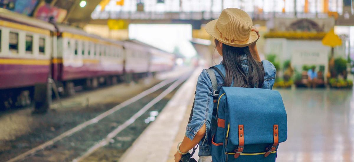 Aufnahme einer Bahnhofshalle. Links sind Gleise zu sehen sowie ein Zug. Rechts im Vordergrund eine junge Frau von hinten, die auf dem Bahnsteig steht und eine Fotokamera in der Hand hält.