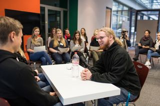 Foto: im Vordergrund sitzen zwei junge Männer an einem Tisch und diskutieren, drumherum sitzen Zuhörerinnen