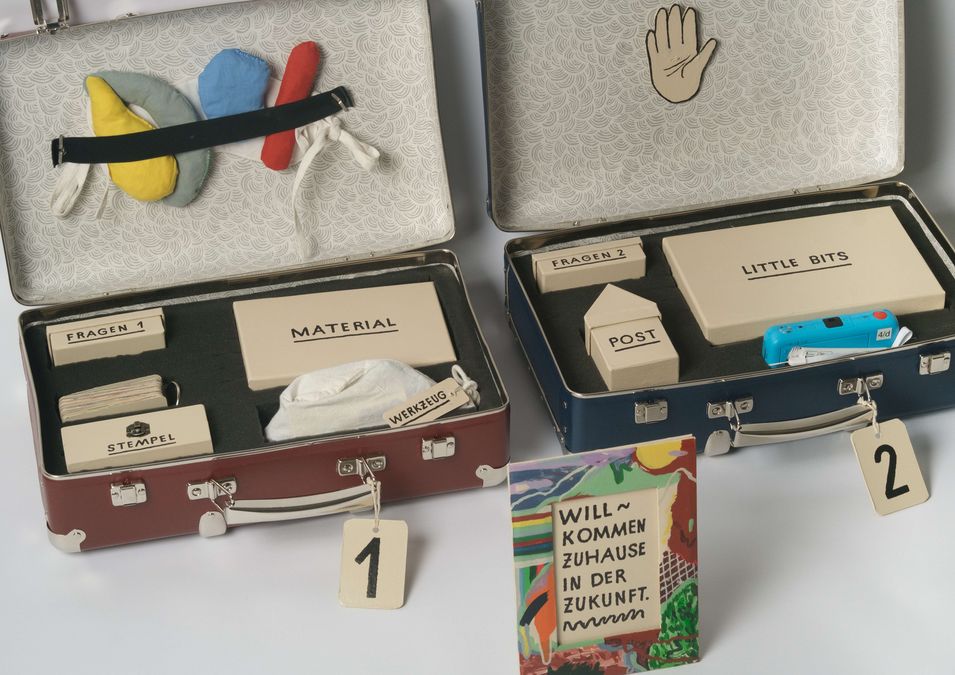 enlarge the image: You can see two open suitcases with numbers on their holders. Inside the suitcases are various envelopes labelled ‘Material’, ‘Little Boxes’ and ‘Stamp’. In front of the suitcases is a colourfully painted picture frame. Inside is the text ‘Welcome home to the future.’