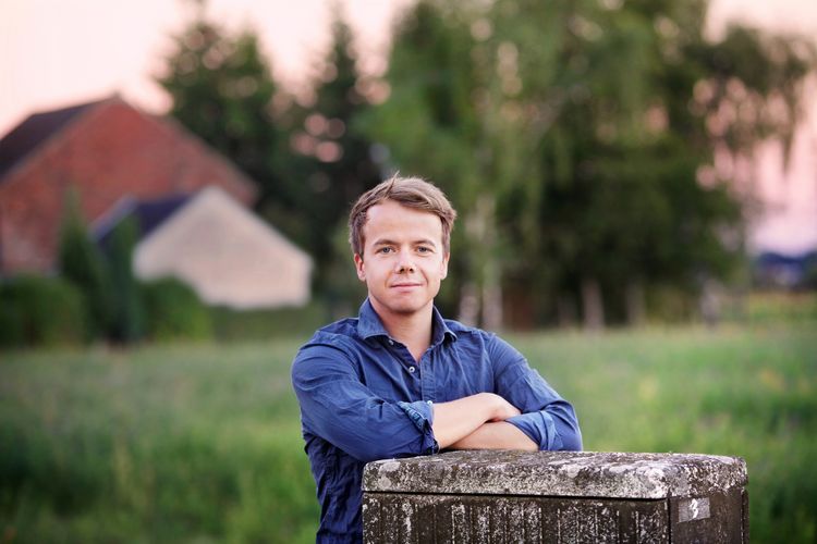 Farbforto von Cornelius Pollmer stehend in der Natur vor einer Wiese mit verschwommenen Häusern im Hintergrund