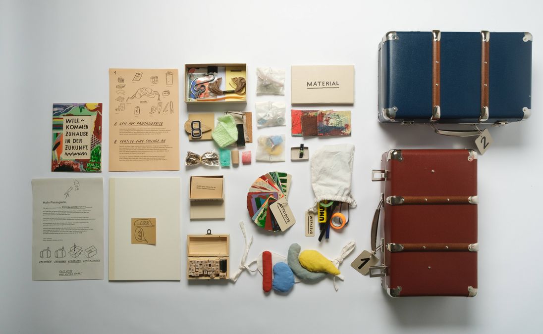 enlarge the image: You can see two open suitcases with numbers on their holders. Inside the suitcases are various envelopes labelled ‘Material’, ‘Little Boxes’ and ‘Stamp’. In front of the suitcases is a colourfully painted picture frame. Inside is the text ‘Welcome home to the future.’