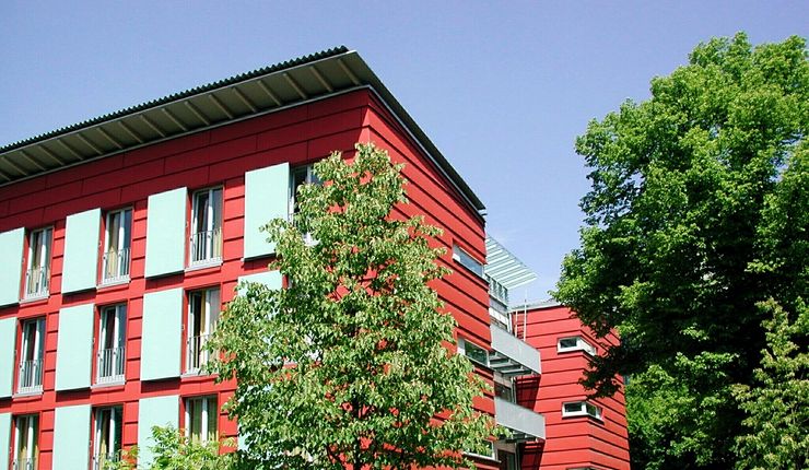 Rote Fassade des Werner-Heisenberg-Hauses der Universität. Die Bäume vor dem Gebäude lassen erahnen, dass es in einer besonders grünen Umgebung steht.