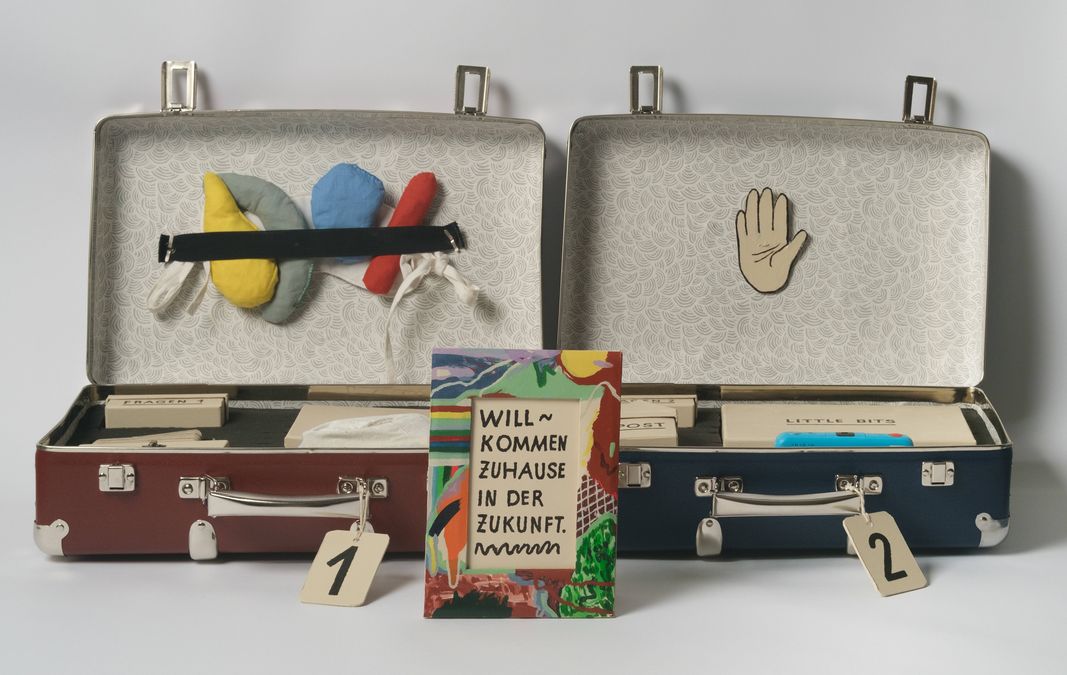 enlarge the image: You can see two open suitcases with numbers on their holders. Inside the suitcases are various envelopes labelled ‘Material’, ‘Little Boxes’ and ‘Stamp’. In front of the suitcases is a colourfully painted picture frame. Inside is the text ‘Welcome home to the future.’