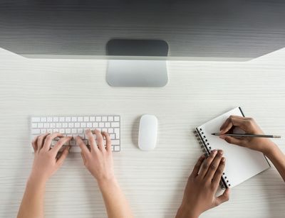 Blick von oben auf einen Computerbildschirm mit Tastatur und einen Notizblock. Am unteren Bildrand erscheinen Arme, die die Tastatur bedienen und auf dem Block schreiben.