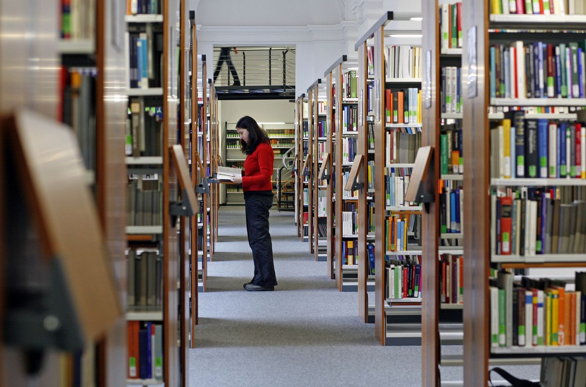 zur Vergrößerungsansicht des Bildes: Mädchen, das ein Buch liest, während es in der Mitte eines Flurs steht, der von Bücherregalen in der Bibliothek umgeben ist.