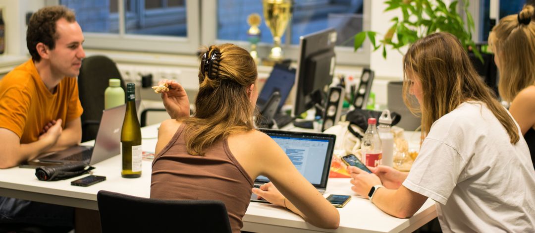 Vier Mitglieder des Fachschaftsrates sitzen mit dem Rücken zur Kamera um einen Tisch.