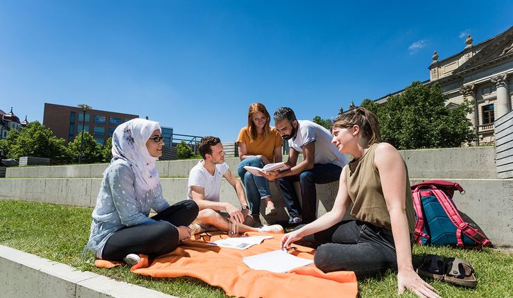 Internationale Studierende sitzen auf einer Picknickdecke in der Sonne vor dem Campus Beethovenstraße 