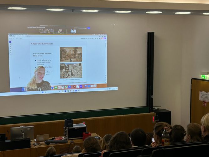 The image shows a screen projection of Janina Dill in front of her presentation, on the wall in the front of a lecture hall filled with students.