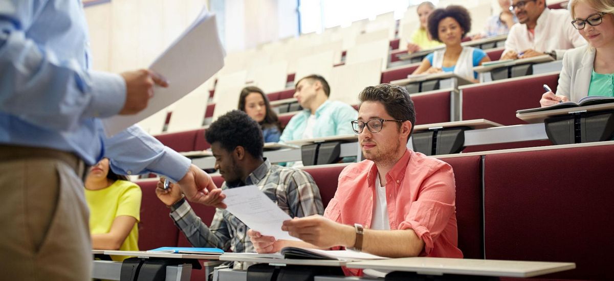 Ein Hörsaal mit Studierenden, der Dozent teilt Unterlagen aus.