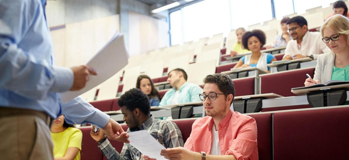 Ein Hörsaal mit Studierenden, der Dozent teilt Unterlagen aus.