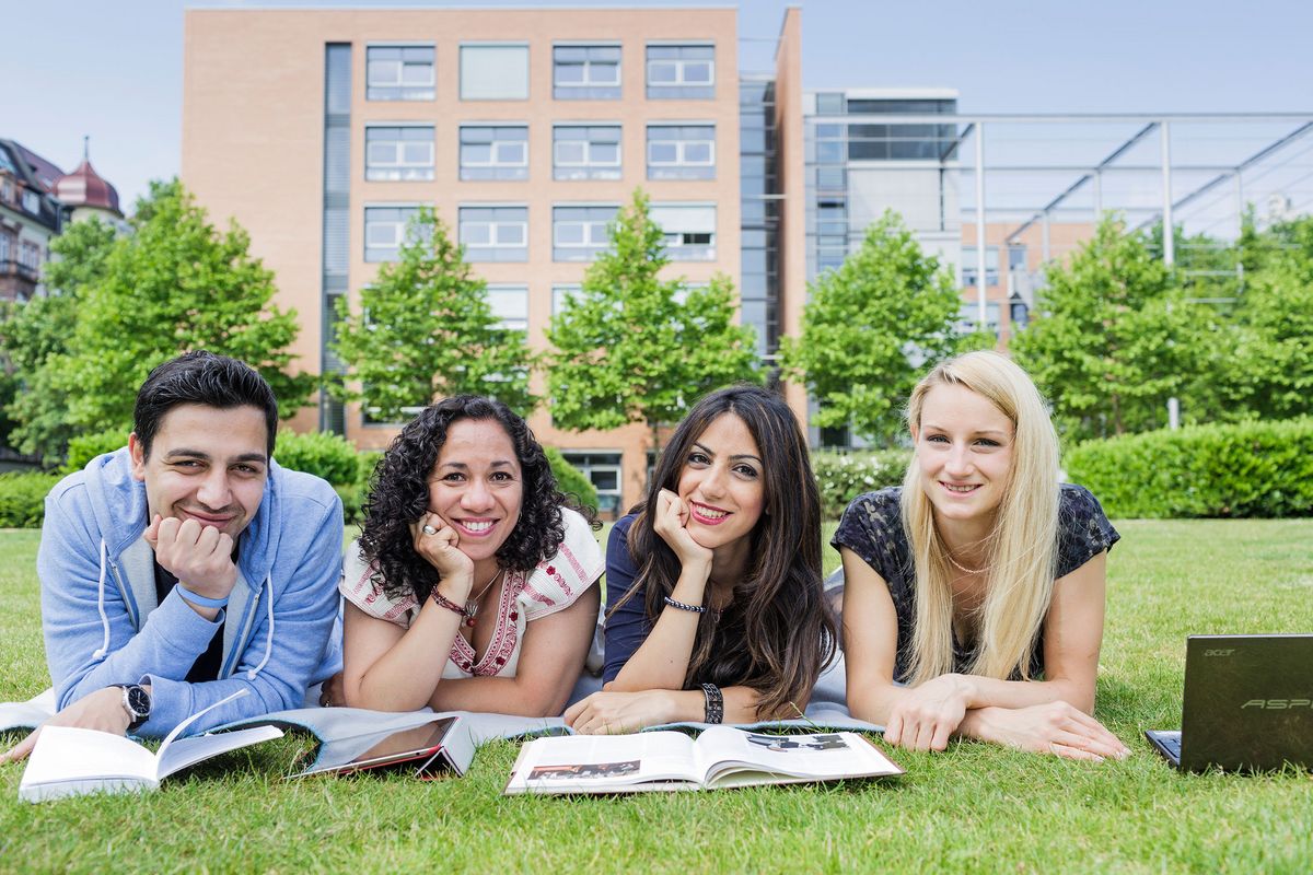 enlarge the image: Foto: vier junge Menschen liegen auf einer Wiese und blicken in die Kamera im Hintergrund ist ein Gebäude zu sehen