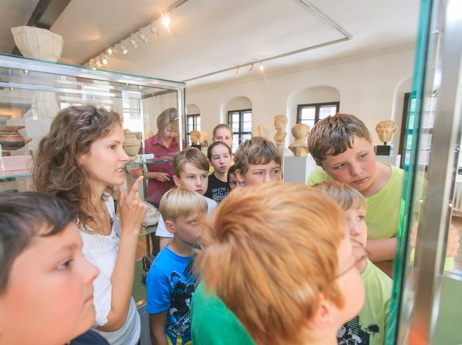 Eine Gruppe Kinder schaut interessiert in eine Vitrine des Antikenmuseums. Der Fokus liegt auf den Gesichtern der Kinder, die Exponte sind nicht abgebildet.